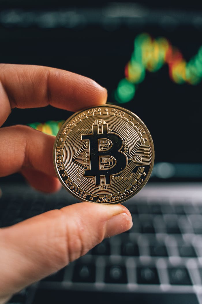 About Close-up of a hand holding a Bitcoin coin in front of a trading screen, symbolizing cryptocurrency trading.